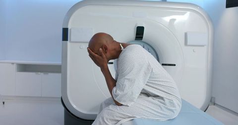 African american man sitting on ct scanner covering face in hospital gown awaiting scan