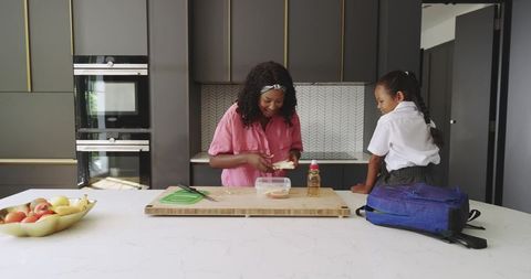 African american mother preparing lunch with schoolgirl on kitchen island, morning routine