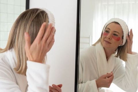Woman applying under-eye gel patches while checking mirror bathroom morning calm routine