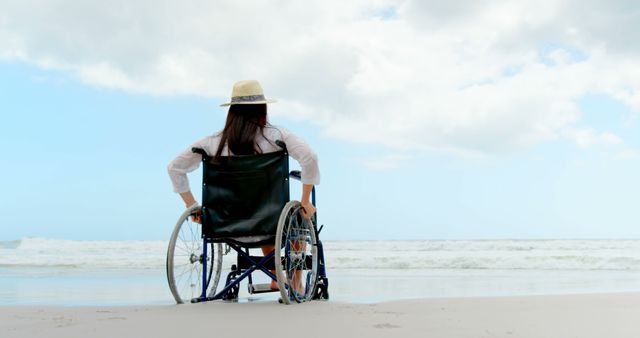 A woman in a wheelchair is facing the ocean waves, wearing a hat, and enjoying the serene beach setting. The sky is partly cloudy, which adds a sense of peacefulness. Ideal for themes such as independence, tranquility, accessibility, and outdoor relaxation.