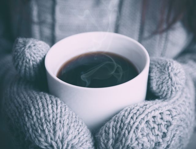 Hands in gray mittens holding a white cup of steaming coffee. Use for winter themes related to staying warm, cozy mornings, or enjoying hot beverages on cold days.
