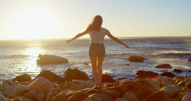Young Caucasian woman enjoys freedom on a rocky beach at sunset. Arms outstretched, she embraces the serene outdoor setting by the sea.