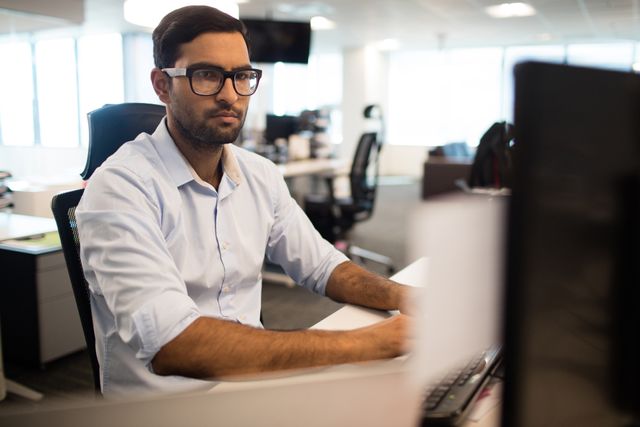 Young businessman concentrating on work at his desk in a modern office. Ideal for use in articles about corporate culture, productivity, office environments, and business technology.