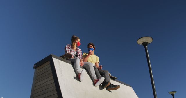Two young friends wearing masks sitting on a ramp outdoors, talking and enjoying the sunny day. Ideal for depicting pandemic safety in public spaces, youth culture, social interactions, and casual conversations among friends while maintaining social distancing.