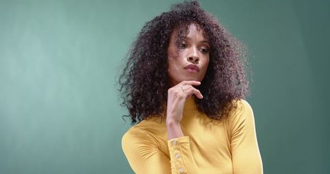 Contemplative woman wearing mustard turtleneck touching chin against soft green backdrop
