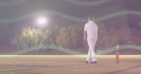 Solitary cricketer walking toward stumps under stadium floodlights with lens flare and empty pitch