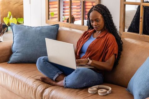 Relaxed african woman working on laptop from sofa, remote work and cozy living room scene