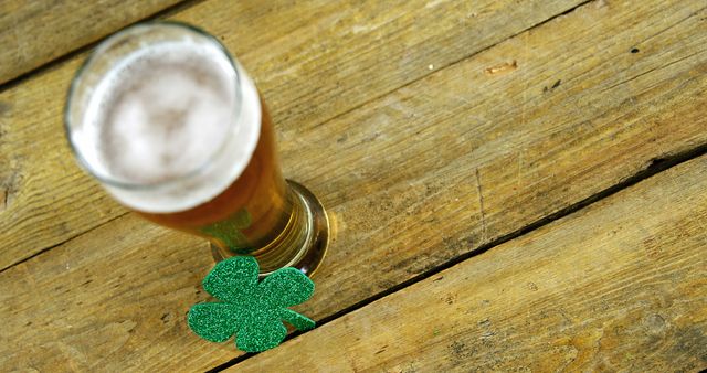 Festive scene with a frothy beer glass placed next to a glittering green shamrock on a rustic wooden table. Perfect for St. Patrick's Day promotions, Irish culture publications, festive advertisements, or celebratory social media posts.