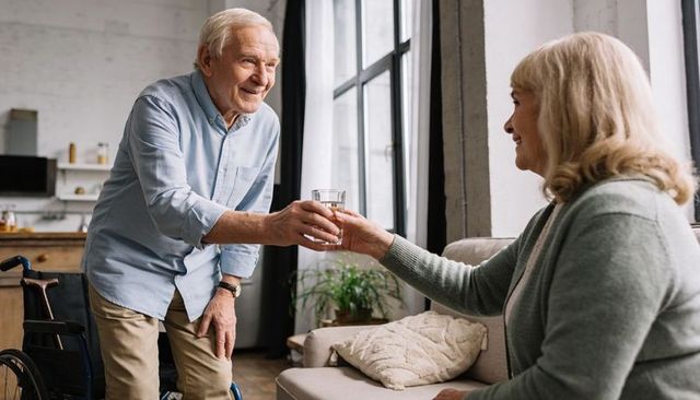 Senior man handing glass of water to smiling partner in cozy sunlit living room with wheelchair