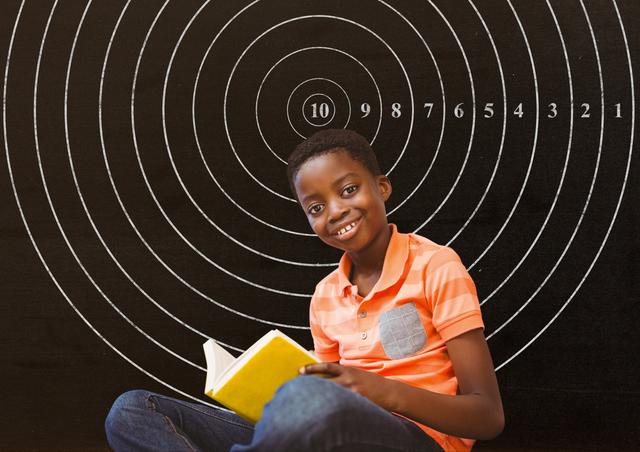Young boy sitting and smiling while holding a notebook in front of a chalkboard with a target design. Ideal for educational content, school promotions, back-to-school campaigns, and childhood learning themes.