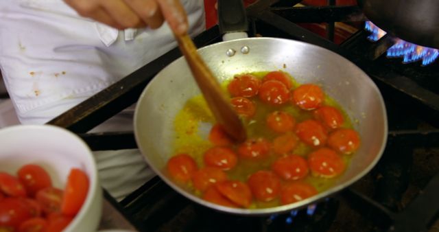 A chef is sauteing halved cherry tomatoes in a frying pan, stirring them over a gas stove. The chef wears white kitchen attire and uses a wooden spatula. This imagery is perfect for illustrating professional cooking techniques, restaurant kitchens, culinary presentations, cooking tutorials, or ingredients and recipe blogs.