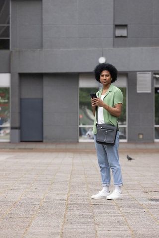 African man standing and using smartphone in urban plaza with crossbody bag