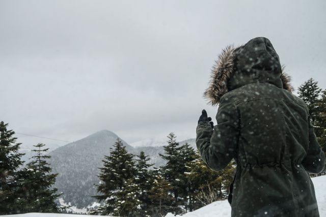 Woman dressed in warm winter parka standing outdoors in snowy mountain landscape. Tall evergreen trees with snow-covered ground and distant mountains enhance the sense of adventure and exploration. This image is perfect for use in travel brochures, winter tourism campaigns, outdoors and nature blogs, advertisements for winter clothing, or holiday cards.