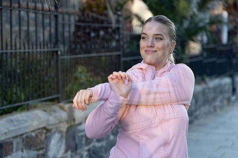 Young woman stretching arms on urban sidewalk wearing pink jacket morning fitness routine