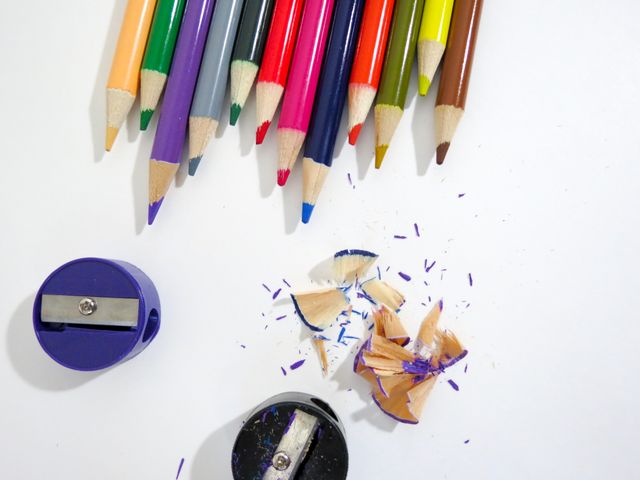 Brightly colored pencils arranged on a white surface alongside pencil sharpeners and wood shavings. Suitable for educational materials, creative workshops, art supply promotions, and student learning contexts. The vibrant colors and scattered shavings evoke creativity and artistic expression.