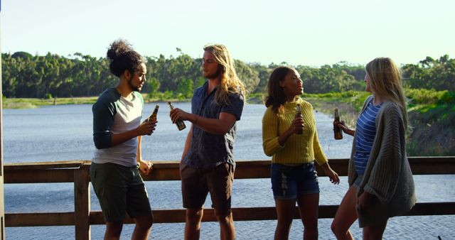 Image showing a group of friends standing on a wooden deck by a scenic lake, enjoying beverages. The diverse group is dressed in casual summer clothing and appears relaxed and cheerful. Suitable for use in advertisements or promotional materials for beverages, outdoor activities, vacations, and social gatherings.