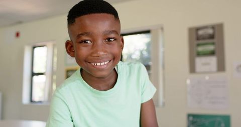 Smiling african boy leaning on desk in bright classroom wearing mint t-shirt school student portrait