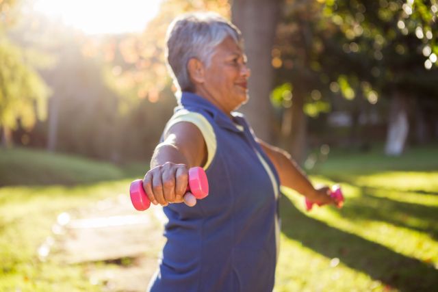 Mature woman lifting pink dumbbells outdoors in a park on a sunny day. Ideal for promoting healthy lifestyles, senior fitness programs, outdoor exercise routines, and wellness campaigns. Can be used in articles, advertisements, and social media posts related to fitness and health for older adults.