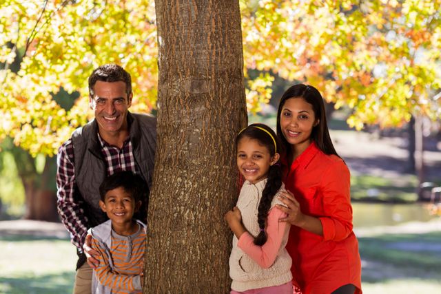 Family of four enjoying time together in a park during autumn. Parents and children smiling and posing by a tree with colorful fall foliage in the background. Ideal for use in advertisements, family-oriented content, lifestyle blogs, and promotional materials for parks or outdoor activities.