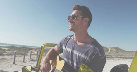 Playing guitar in convertible on coastal dunes wearing aviator sunglasses and tie-dye tee