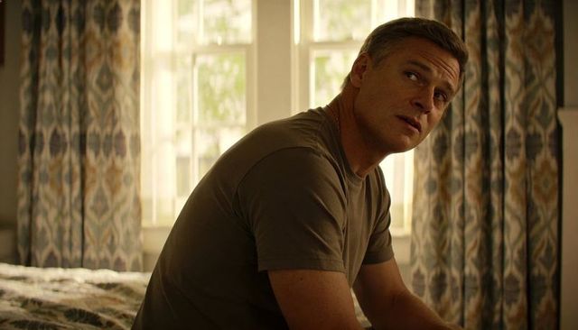 Contemplative man sitting on bed in olive tee backlit by window with patterned drapes