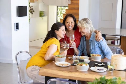 Multicultural mature women laughing, hugging, toasting white wine at modern dining table