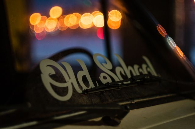 Closeup of a vintage car's windshield featuring a stylish Old School sticker with out-of-focus bokeh city lights in the background. Perfect for use in content related to automotive culture, retro themes, urban photography, night scenes, and nostalgia.