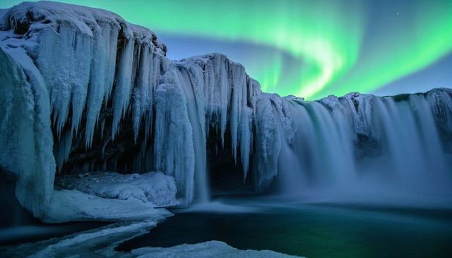 Aurora dancing over frozen waterfall with dramatic icicles and misty plunge pool