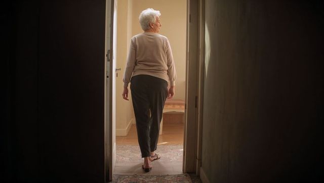 Senior woman walking from dim hallway into sunlit room, back view, aging at-home lifestyle