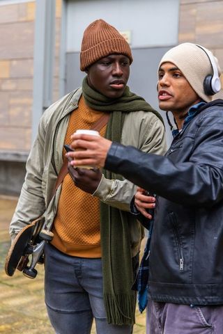 Two diverse young men chatting while holding skateboard and coffee in urban streetstyle