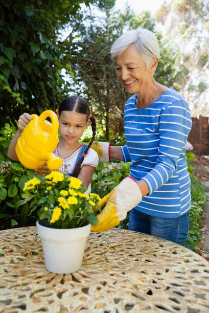 Smiling senior woman standing by granddaughter watering yellow flowers on table at backyard