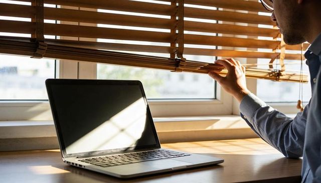 Man adjusting wooden blinds at home office desk with laptop glare, soft morning sunlight