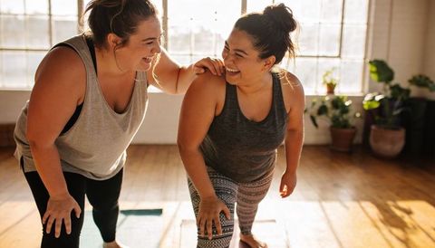 Laughing women stretching together in sunlit yoga studio showing supportive fitness bond