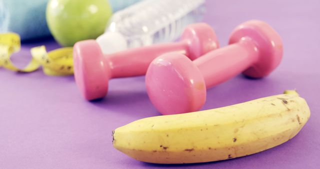 A banana, pink dumbbells, a water bottle, and a measuring tape are arranged on a purple surface, symbolizing health and fitness. These items represent a balanced approach to wellness, combining nutrition, hydration, and exercise.