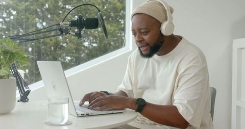 African man hosting sunlit home podcast with headphones and laptop in minimalist workspace