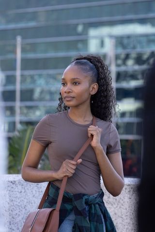Confident young woman standing in city plaza holding leather crossbody bag, plaid shirt tied waist
