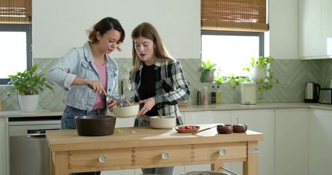 Diverse women cooking together in sunlit modern kitchen, scooping pasta into ceramic bowls