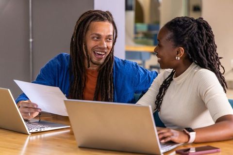 Diverse coworkers collaborating at laptops, smiling and reviewing document in office
