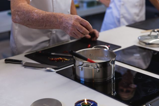 Mid section of male chef cooking in a modern busy kitchen, adding ingredients to a pan on electric hob. Cookery class at a restaurant kitchen. Workshop cooking food.