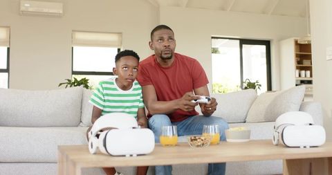 African american father and son playing video games on couch with vr headsets and controller