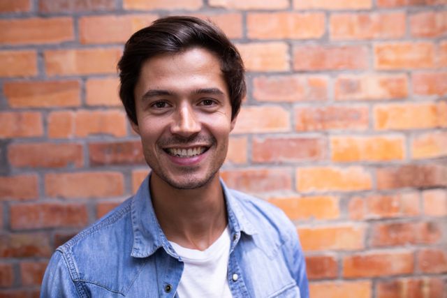 Portrait of a confident happy Caucasian businessman with short dark hair working in a modern creative office, wearing a denim shirt smiling and looking at camera, brick wall in the background.