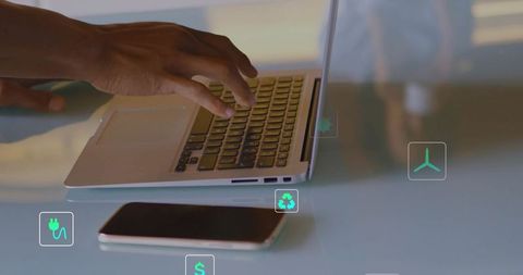Right hand typing on aluminum laptop with smartphone and floating eco icons on glass desk