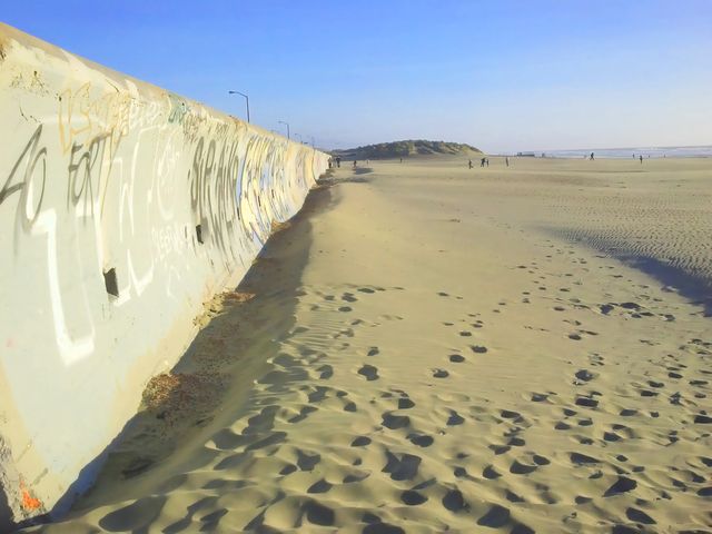 Sandy beach stretches into distance with a graffiti-covered wall on one side. Clear blue sky overhead, footprints scattered across sand. People walking in distance near sand dunes. Ideal for depicting seaside leisure, urban art, outdoor activities, coastal landscapes.