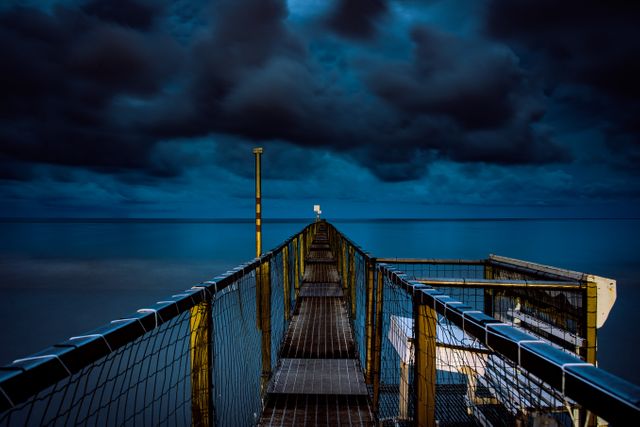 Dramatic scene of a pier extending into the distance under a stormy night sky. Dark, thick clouds create an ominous and moody atmosphere, contrasting with the calm water below. Ideal for use in creative projects depicting solitude, adventurous journeys, or the beauty of nature. Suitable for backgrounds, travel blogs, or calming inspirational imagery.