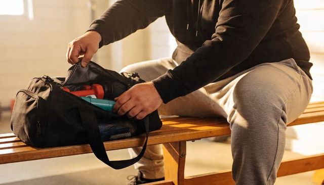 Man packing gym duffel on bench preparing workout with shaker bottle and dumbbells