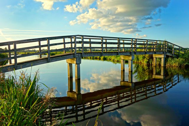 This image captures a wooden bridge spanning over a calm river during sunset, reflecting the picturesque clouds and surrounding greenery. Perfect for projects related to nature, travel, tranquility, outdoor exploration, and scenic beauty.