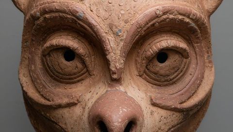 Weathered terracotta owl mask close-up highlighting textured clay, hollow eyes and prominent beak
