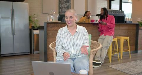Smiling woman with shaved head working on laptop in cozy cafe during virtual meeting