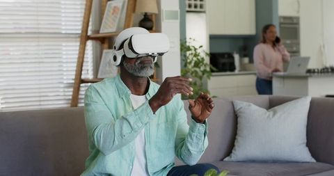 Man exploring virtual reality at home with VR headset while partner working in kitchen