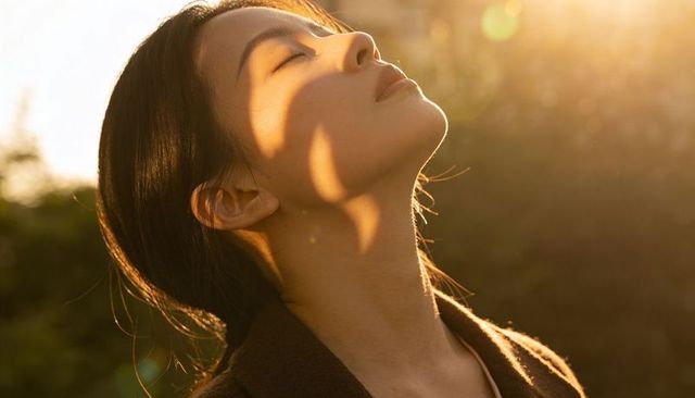 Asian woman basking in golden hour light tilting head back with closed eyes, tranquil